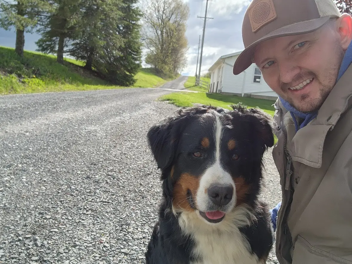 Selfie of a man in a cap beside a Bernese Mountain Dog on a gravel rural road with a white house in the background.