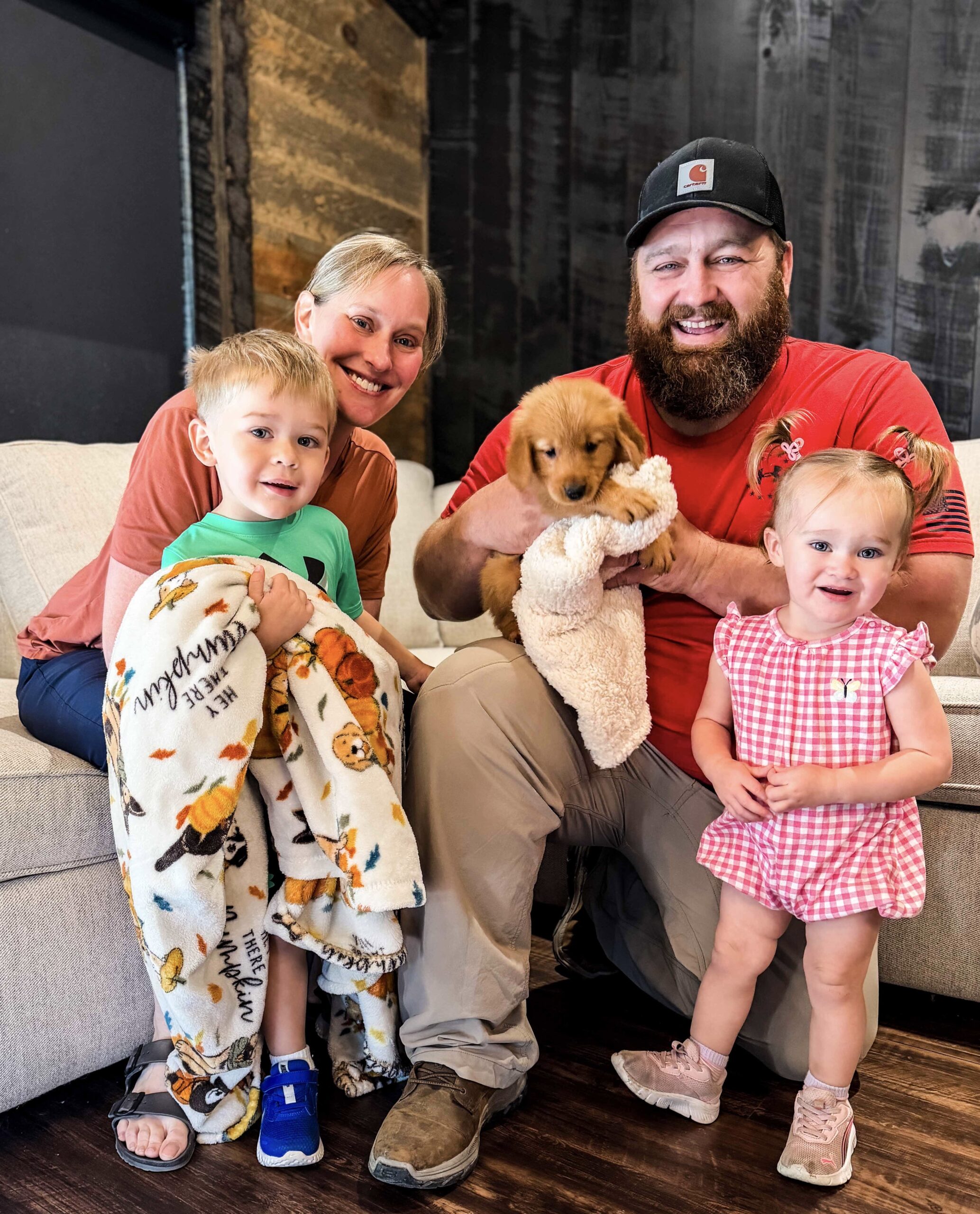 Smiling family of four on a couch with a small golden puppy and a blanket in the man's arms.